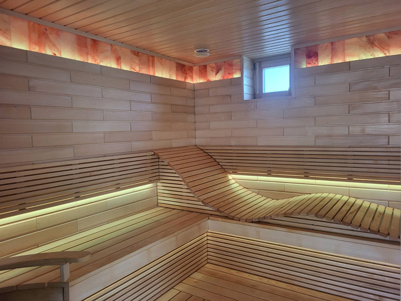 Sauna in legno chiaro con panca ondulata e illuminazione a LED.