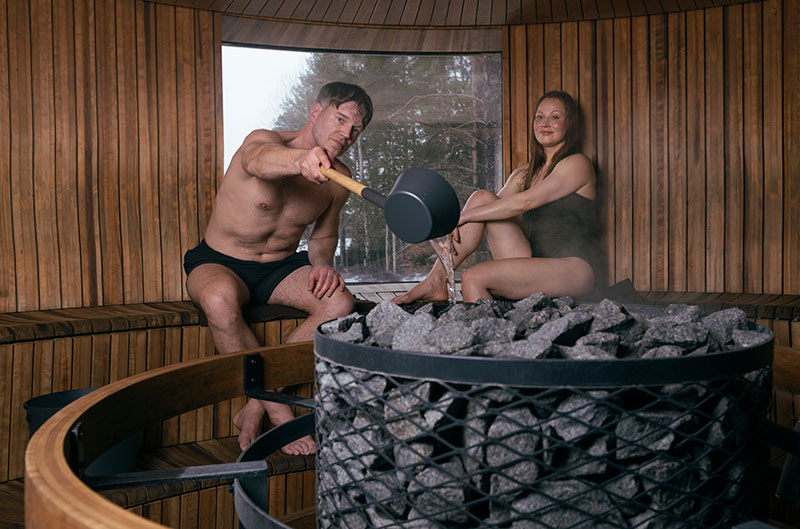 Un uomo versa l'acqua sulle pietre riscaldate della sauna con un mestolo, una donna è seduta lì vicino; una sauna di legno rotonda e piena di vapore, con una finestra.